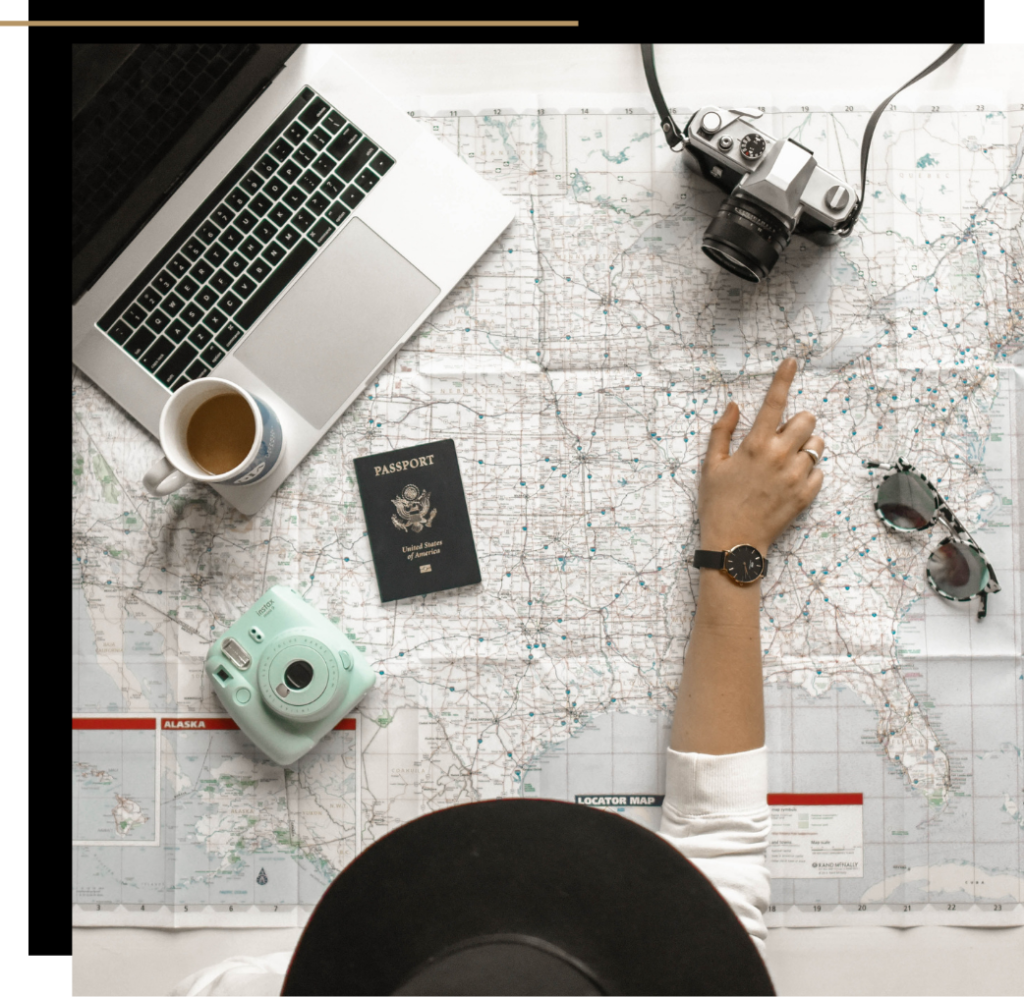 A girl in a hat pointing towards a camera with a map, laptop, passport and sunglasses